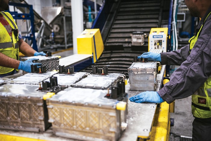 Two workers wearing blue gloves stand over a table holding large batteries. A conveyor belt is attached to the table.