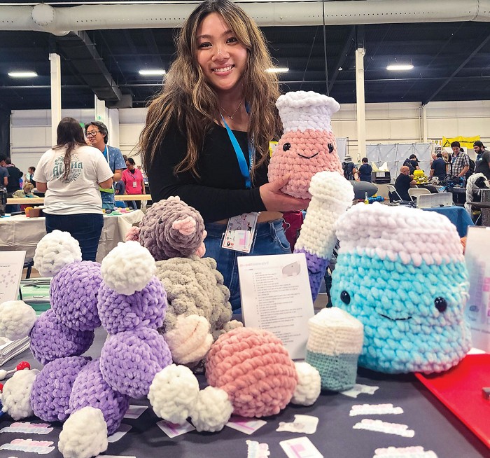 A woman holds a crocheted Erlenmeyer flask plushie. She stands behind a table that has other chemistry-related plushies on it, including a beaker, a benzene molecule, a water molecule, and a mole.