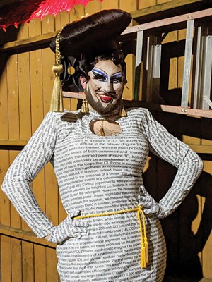 A bearded drag queen poses for the camera. She is wearing a hat that resembles a graduation cap and a white dress with black text written all over it.