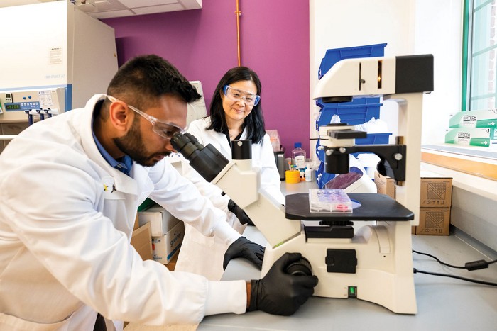 One scientist looks into a microscope while another observes. Both are wearing lab coats, black nitrile gloves, and safety glasses.