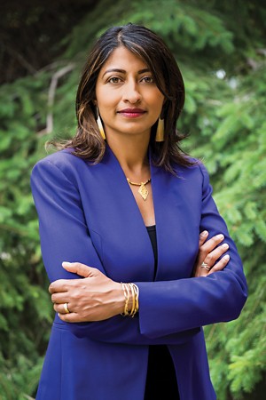 A woman in a blue suit stands with her arms crossed, lightly smiling at the camera.