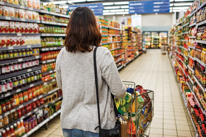A shopper pushes a cart full of groceries down an aisle of a store stocked full of processed food.
