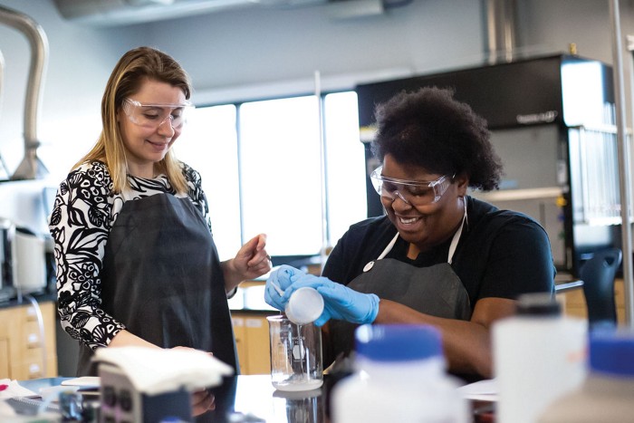 A standing woman (left) wearing an apron and safety glasses looks on while a seated student adds a white powder to a beaker on a lab bench.