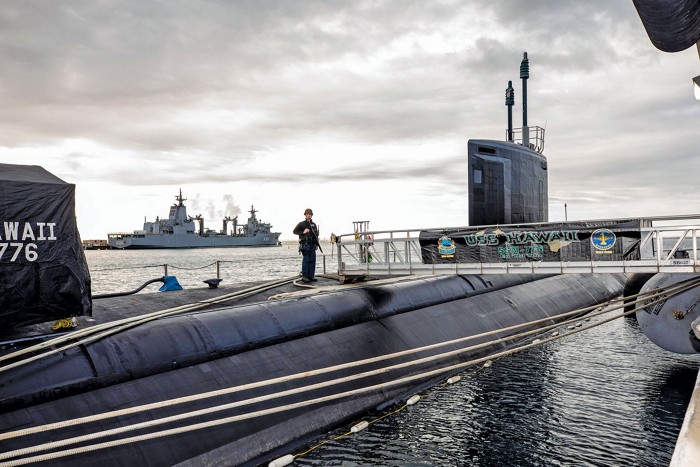 A US Navy submarine sitting in a dock with a sailor on top standing watch.