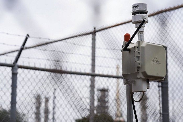 An air monitor on a pole in front of a fence topped by barbed wire.