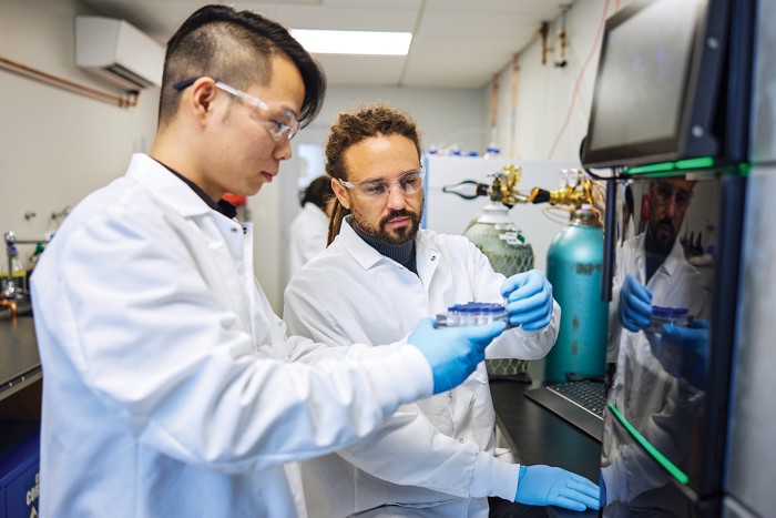 Two scientists wearing lab coats, safety glasses, and nitrile gloves examine a tray in a laboratory setting.