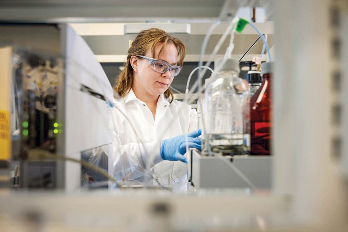A scientist wearing a lab coat, safety glasses, and nitrile gloves is working among various glass containers and tubes.