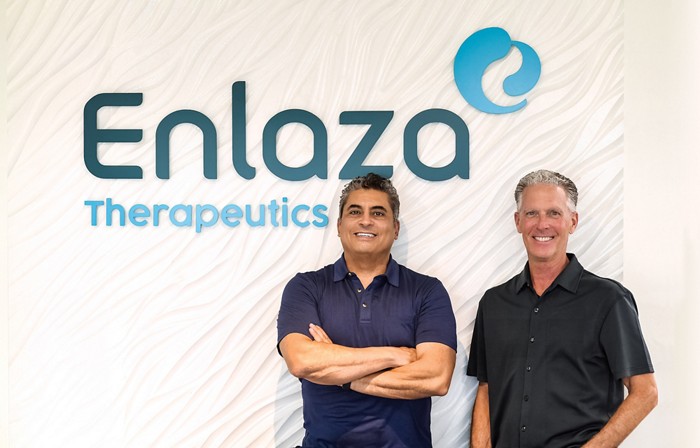 Two men stand in front of a wall with the Enlaza Therapeutics company logo on it.