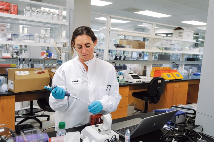 A woman in a lab coat and gloves pipettes from a microcentrifuge tube. She’s standing in front of a NanoDrop spectrophotometer.