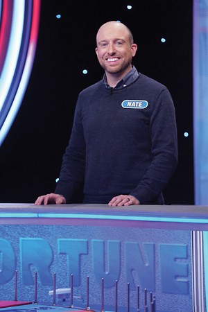 A smiling man in a blue sweater stands in the wheel of fortune set. On his chest is a name bage saying Nate
