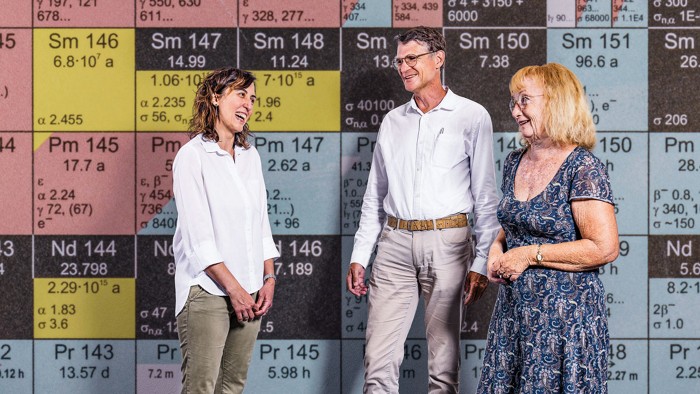 Three people stand in front of a colorful tiled wall, which appears to show information about isotopes.