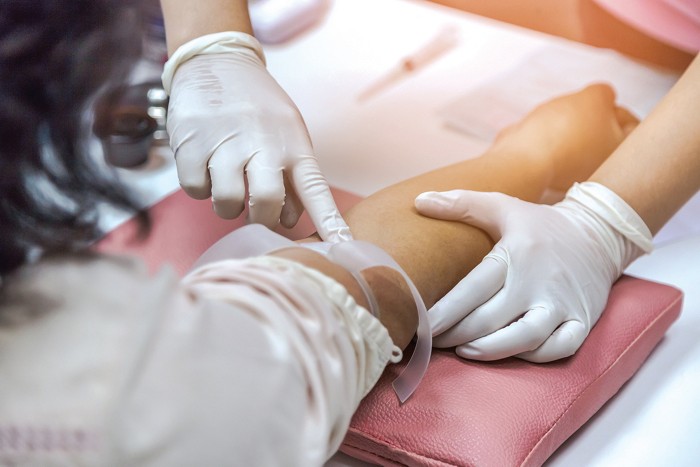 Photo shows a gloved hand pressing against the inner elbow of a person whose arm has a tourniquet above the elbow.