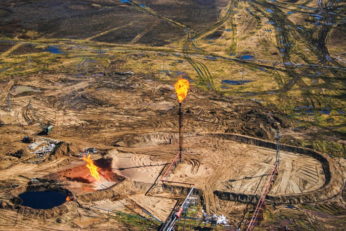 An aerial view of a dirt field of with oil drilling equipment on it. A large flame emanates from a tower in the middle of the field.