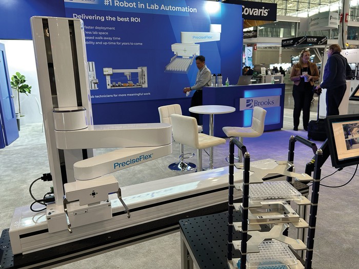 A robotic arm is displayed at a booth sponsored by Brooks Automation. It is mounted on a platform that allows it to move back and forth on a single axis.