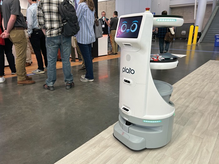 A robot rolls across a showroom floor. It has three shelves mounted on its back. On its front, a screen displays a cartoon smiling face.