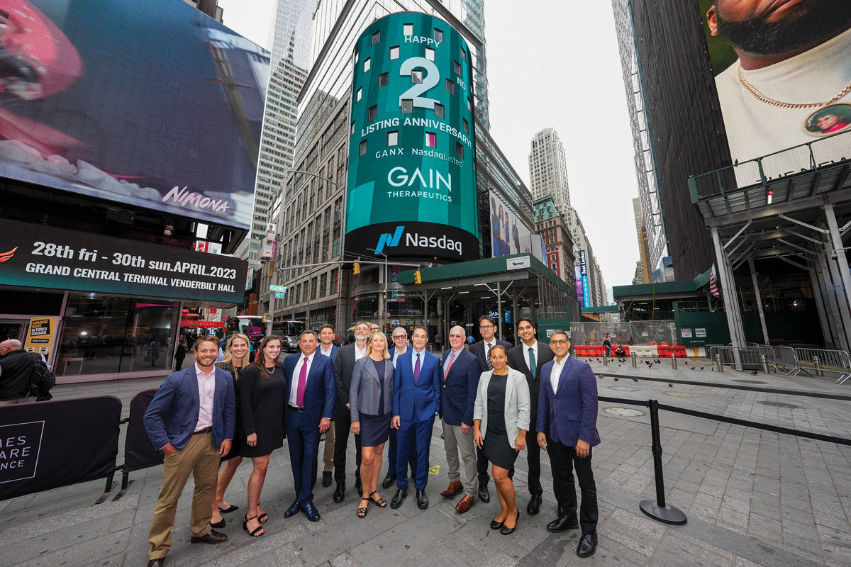 Fourteen people in business formal wear stand in front of the Nasdaq MarketSite building, whose digital marquee displays "Happy 2nd Listing Anniversary Gain Therapeutics."