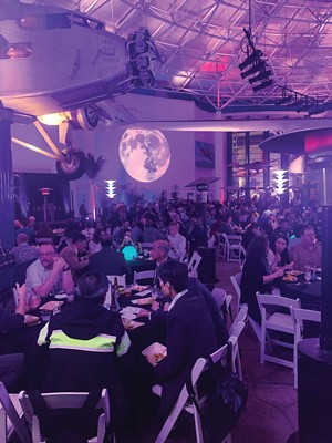 A crowd of people at tables and chairs at a party, bathed in blue light. Above their heads are various aircraft.