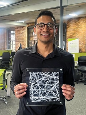 A man in casual clothes holds a framed electron microscope image of a nonwoven fiber mesh.