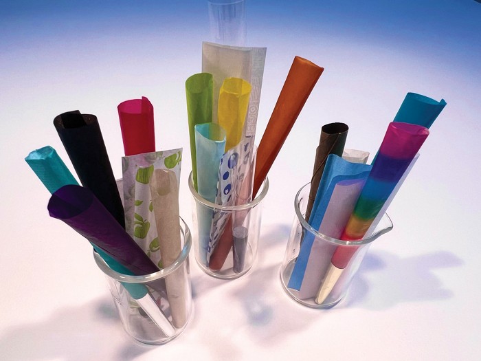 Three beakers contain rolling papers and preformed paper cones for cigarettes or cannabis. The roughly two dozen papers come in a spectrum of colors, including one cone that is striped with a rainbow of colors.