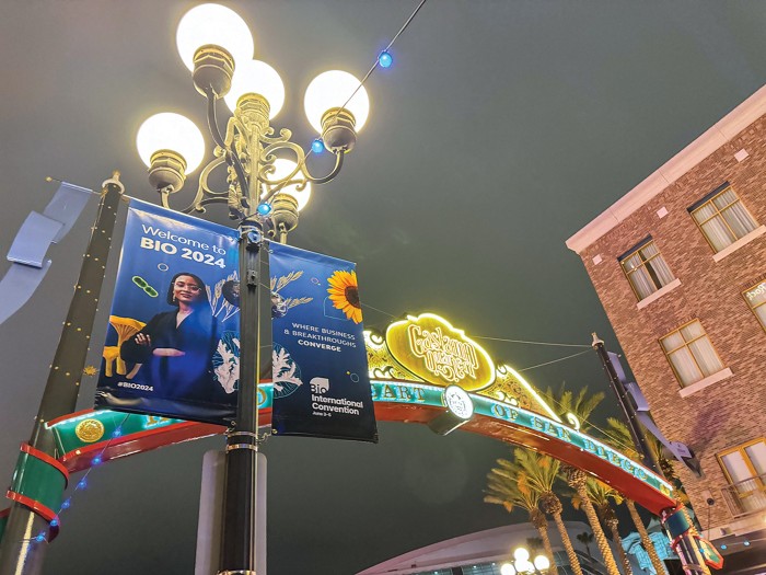 An advertisement for BIO 2024 is displayed across two flags posted up a streetlight. In the background, a pole lantern lights the night sky next to a neon archway that reads "Gaslamp Quarter."