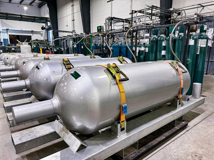 Gas tanks standing up are connected by hoses to much larger gas tanks lying down in a warehouse.