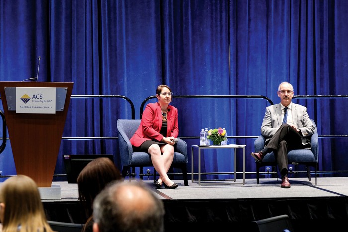 A woman sits in a chair to the left and a man sits in a chair to the right as they look to an audience and answer questions.