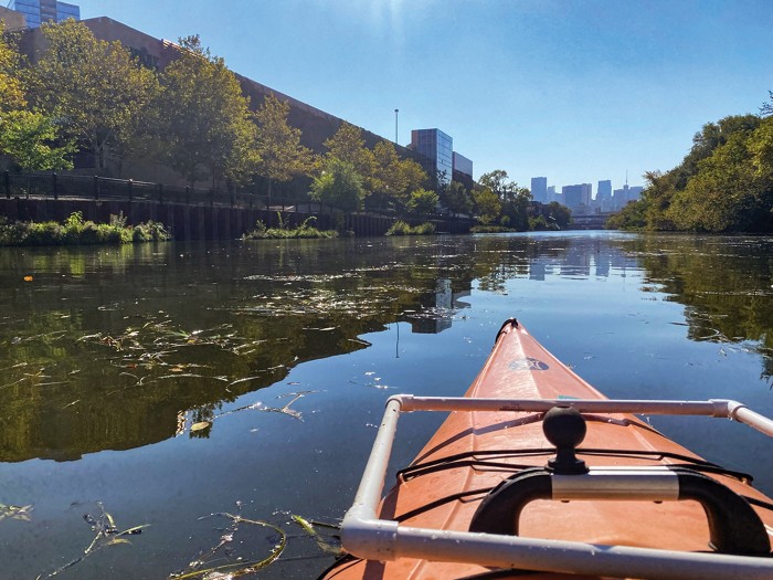 Image of a kayak on a river taken from the kayaker's seat. The bow of the boat is in the foreground, with a square made of pipes lying on the orange kayak. In front of the kayak is a river, an embankment, and a city skyline in the distance.