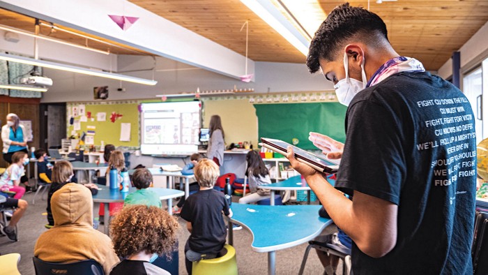 A man wearing a face mask inside a classroom full of students and a teacher. The man is measuring the air quality levels using a device.