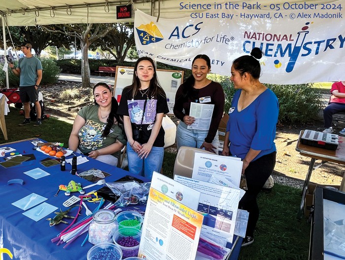 Four adults stand behind a table containing tubs of beads and other items.