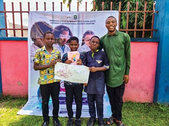 One adult and three children stand in front of an ACS promotional poster and hold a large piece of paper with a purple mark on it.