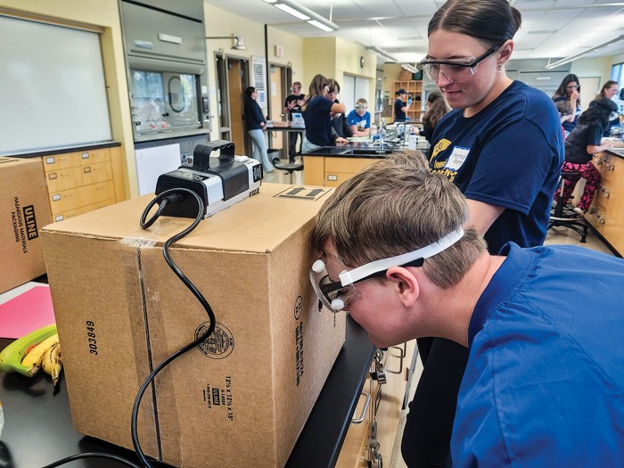 A student in safety goggles looks into a cardboard box with a fluorescent lamp on the top.