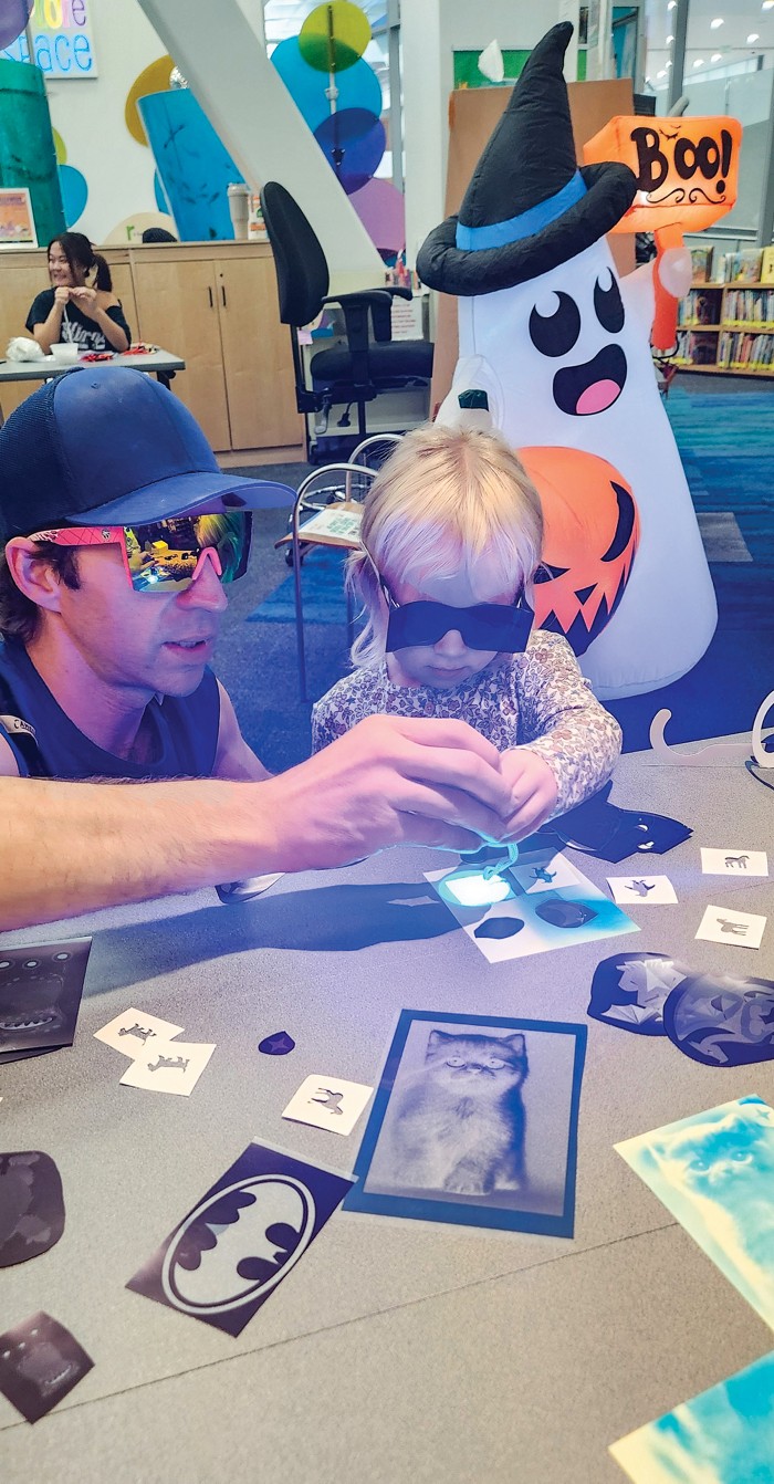 A child and adult wearing safety glasses point an ultraviolet flashlight at a stencil.