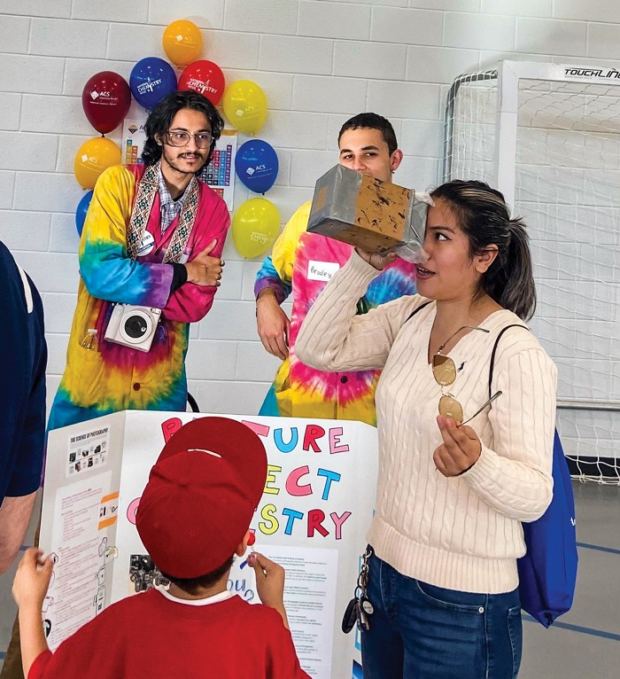 A child and two adult volunteers watch an event attendee look through a pinhole camera.