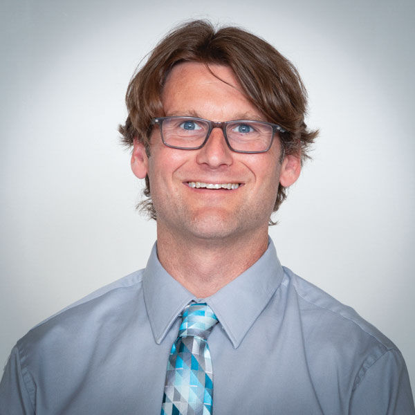 A man with curly brown hair wears glasses, a grayish blue button-down shirt, and a patterned gray-and-blue tie and smiles for an informal portrait shot.