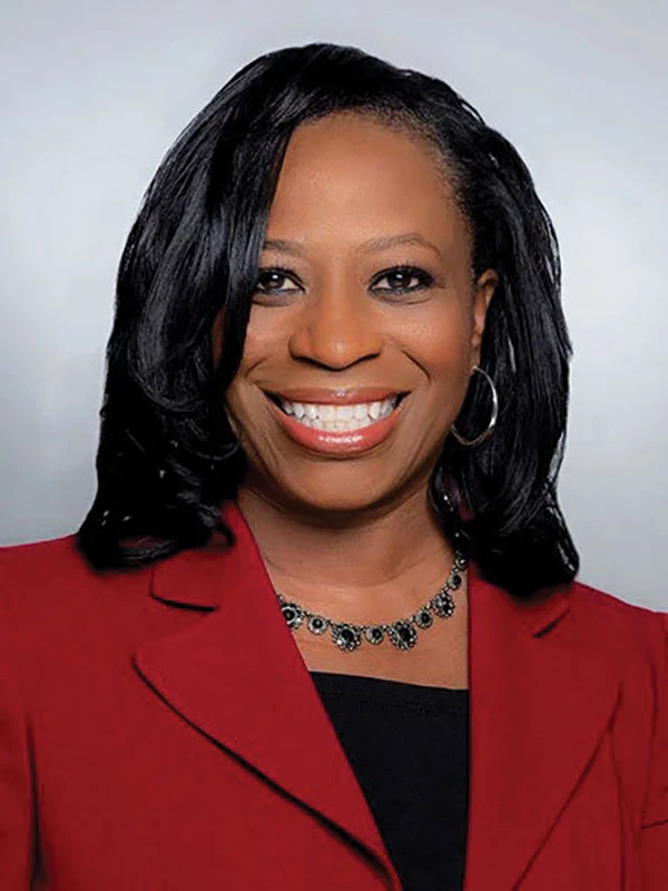 A smiling woman wearing a black shirt and dark red blazer poses for a formal portrait shot.