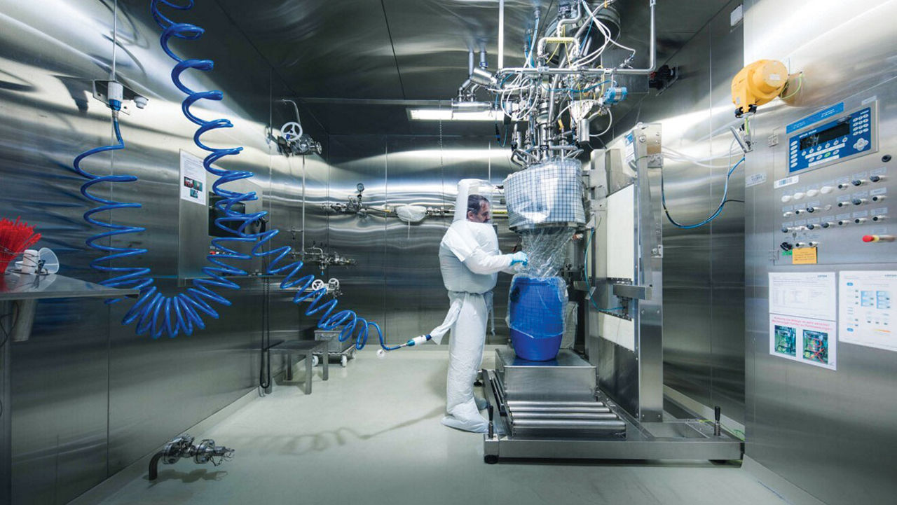 A worker stands in white full-body protective gear in a facility with stainless steel walls.