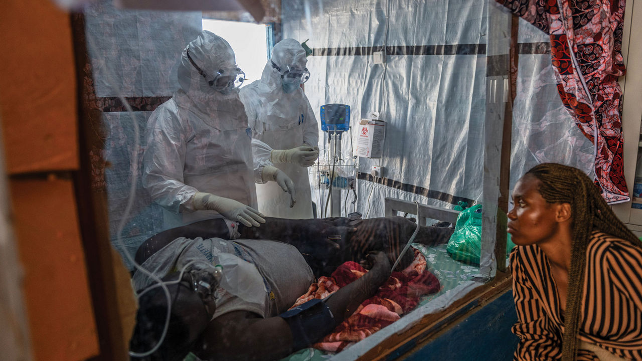 Two health-care workers in full protective wear attend to a patient while a person wearing a striped shirt watches.