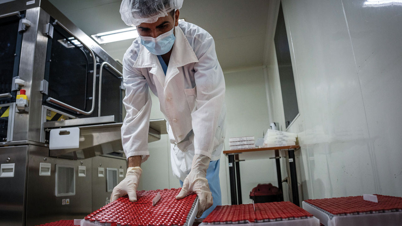 A worker wearing a helmet, gloves, a mask, and a lab coat handles trays of vials with red caps.