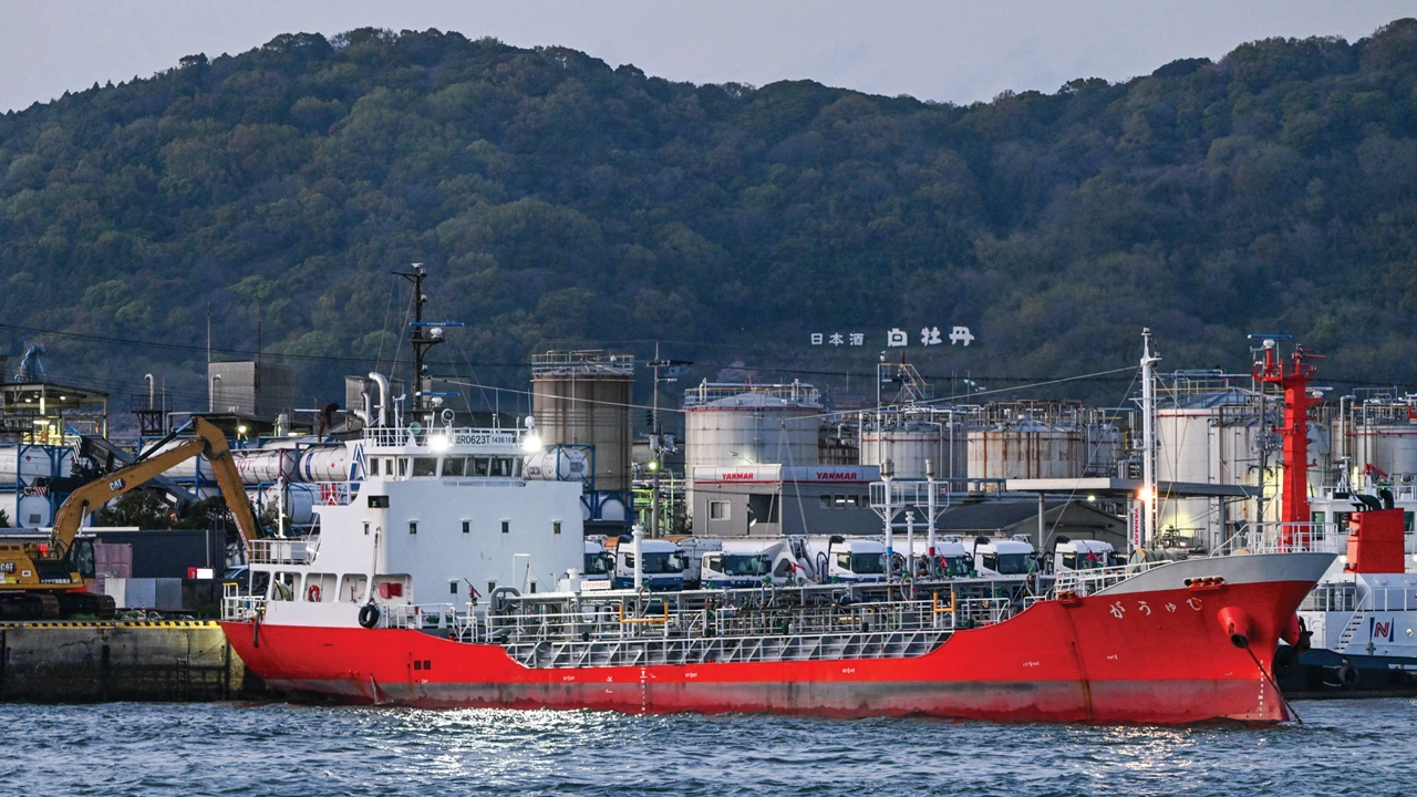 A ship is docked in front of an array of storage tanks, and green hills loom in the background.