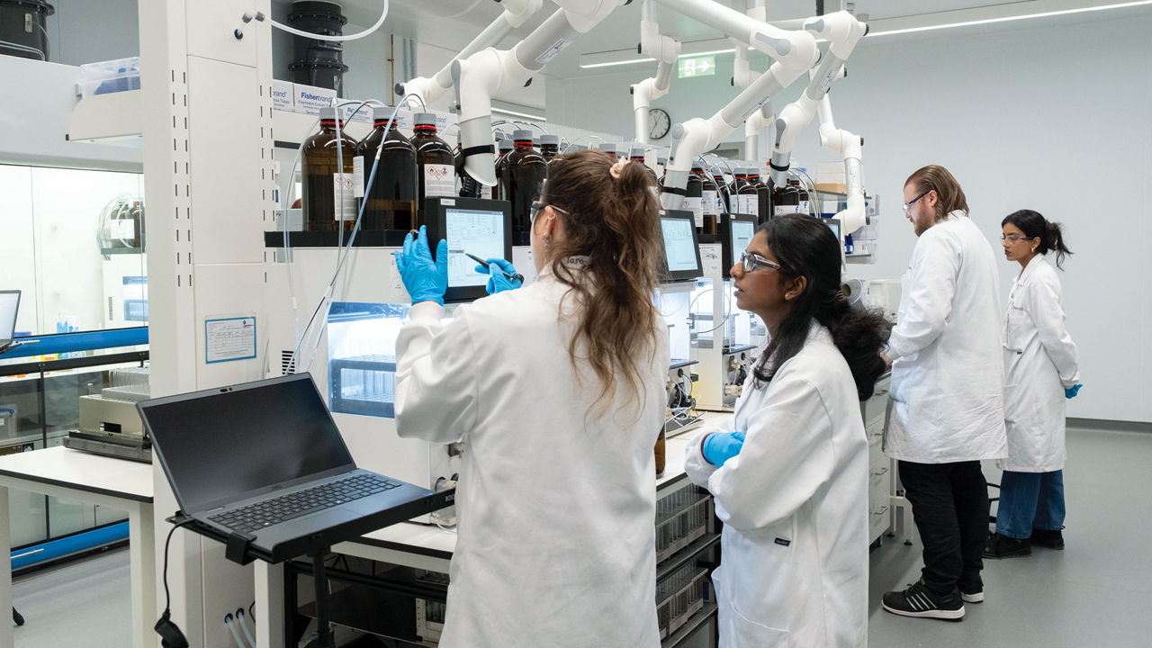 Four scientists wearing white lab coats look at various screens in a laboratory.