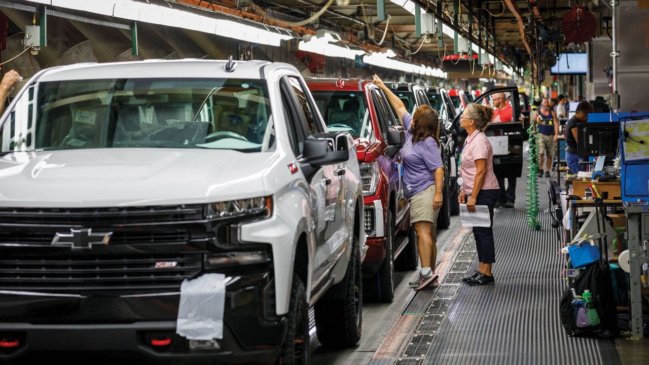 Two workers inspect white and red pickup trucks as they roll down an assembly line.
