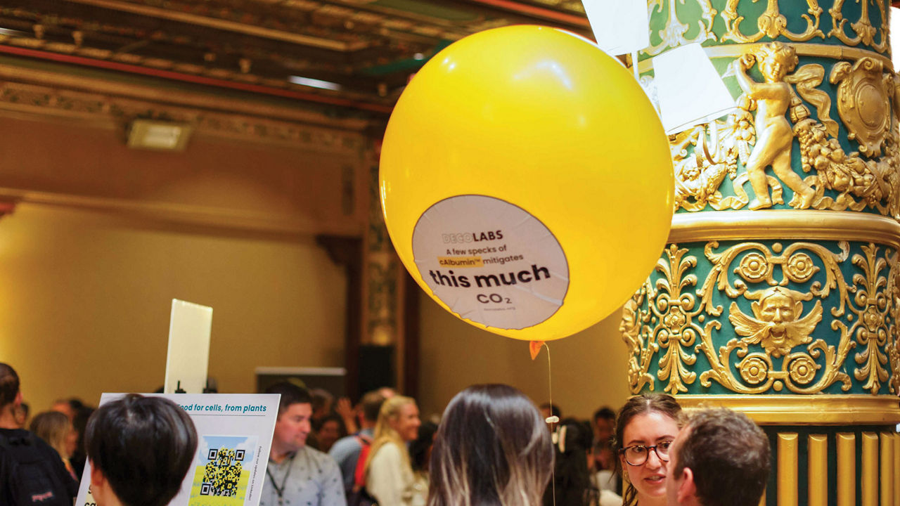 A crowd milling in a ballroom with a large yellow balloon hovering overhead that advertises a low-CO<sub>2</sub> cell culture medium.