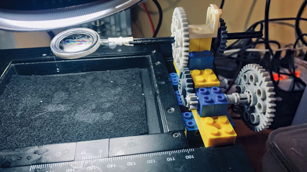 A series of five Lego gears of varying sizes mounted on Lego blocks. Out of one of the gears extends a short rod connected to a plastic loop that contains an iridescent thin film of soapy water.