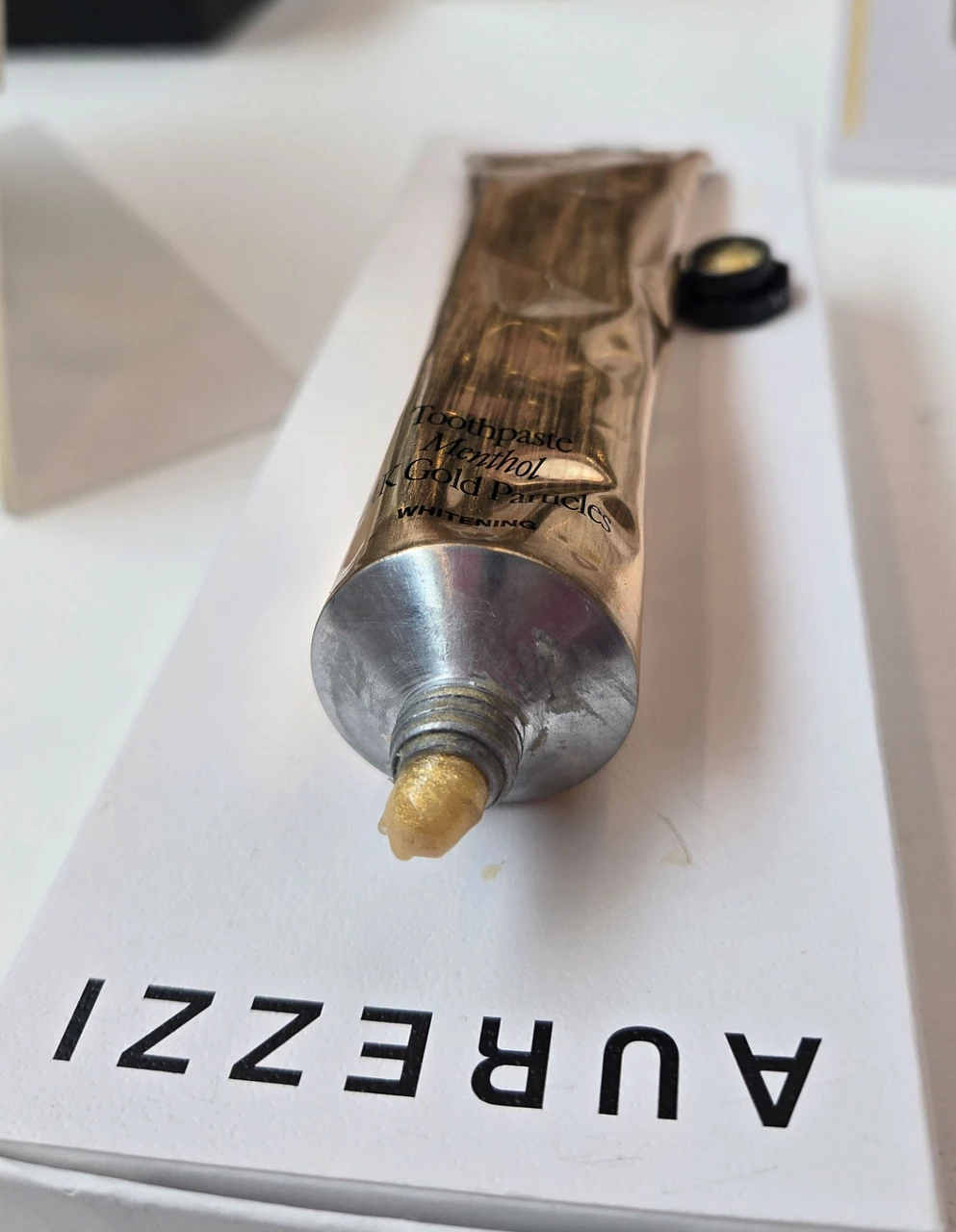 A metal toothpaste tube with a gold-colored paste coming out the spout, with the label AUREZZI beside the tube.
