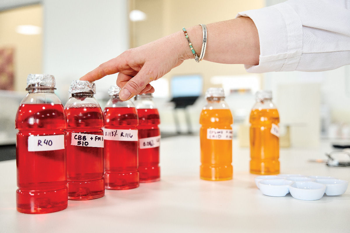 A person points to a red sports drink bottle. Several other bottles of red sports drinks and two orange sports drinks are also on the table. 