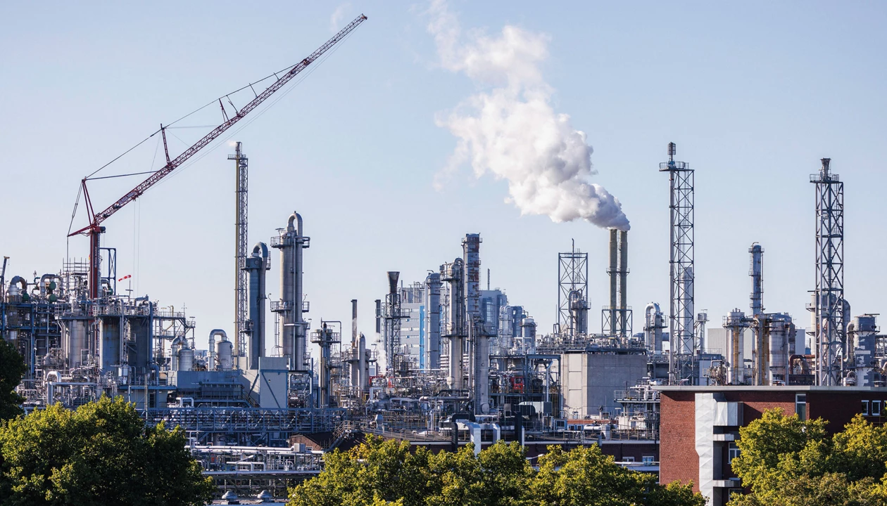 A large chemical plant with multiple buildings and metallic towers stands against a blue sky with trees in the foreground.