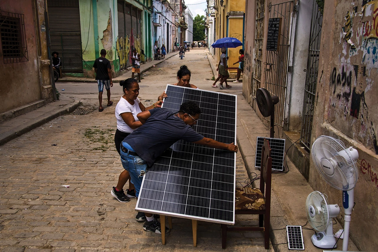 Three people hold a large solar panel on a cobblestone street, with two smaller solar panels and three fans set up on the sidewalk near them. Buildings with graffiti line the street, and people sit, walk, and ride bikes in the distance.