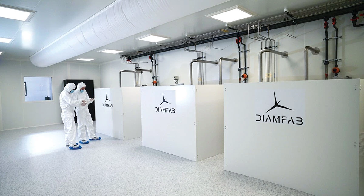 Two men dressed in white overalls look at clipboards in a white pilot production room. Three large boxes fed by pipes say “Diamfab” on the side.