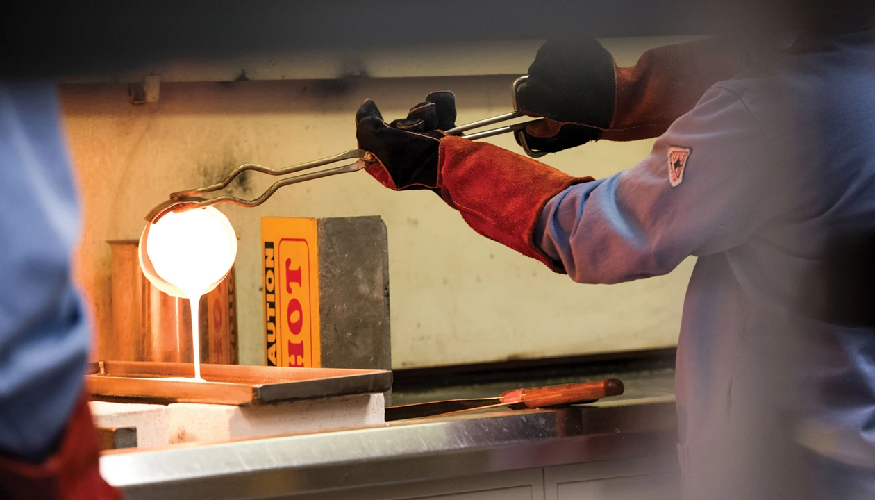 Hands wearing thick safety gloves hold metal tongs holding a container with a molten substance pouring out onto a tray on a counter.