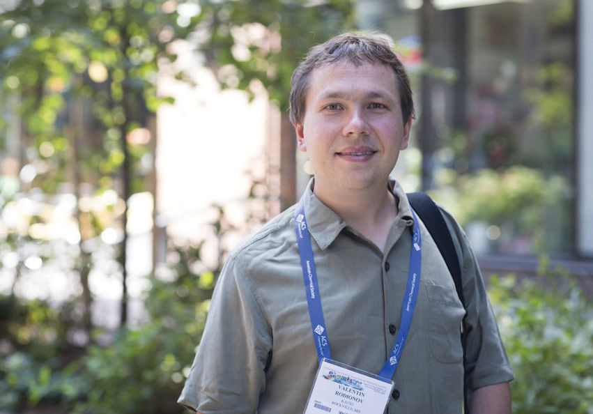 A person wearing a collared shirt and a conference badge poses in front of a leafy background.